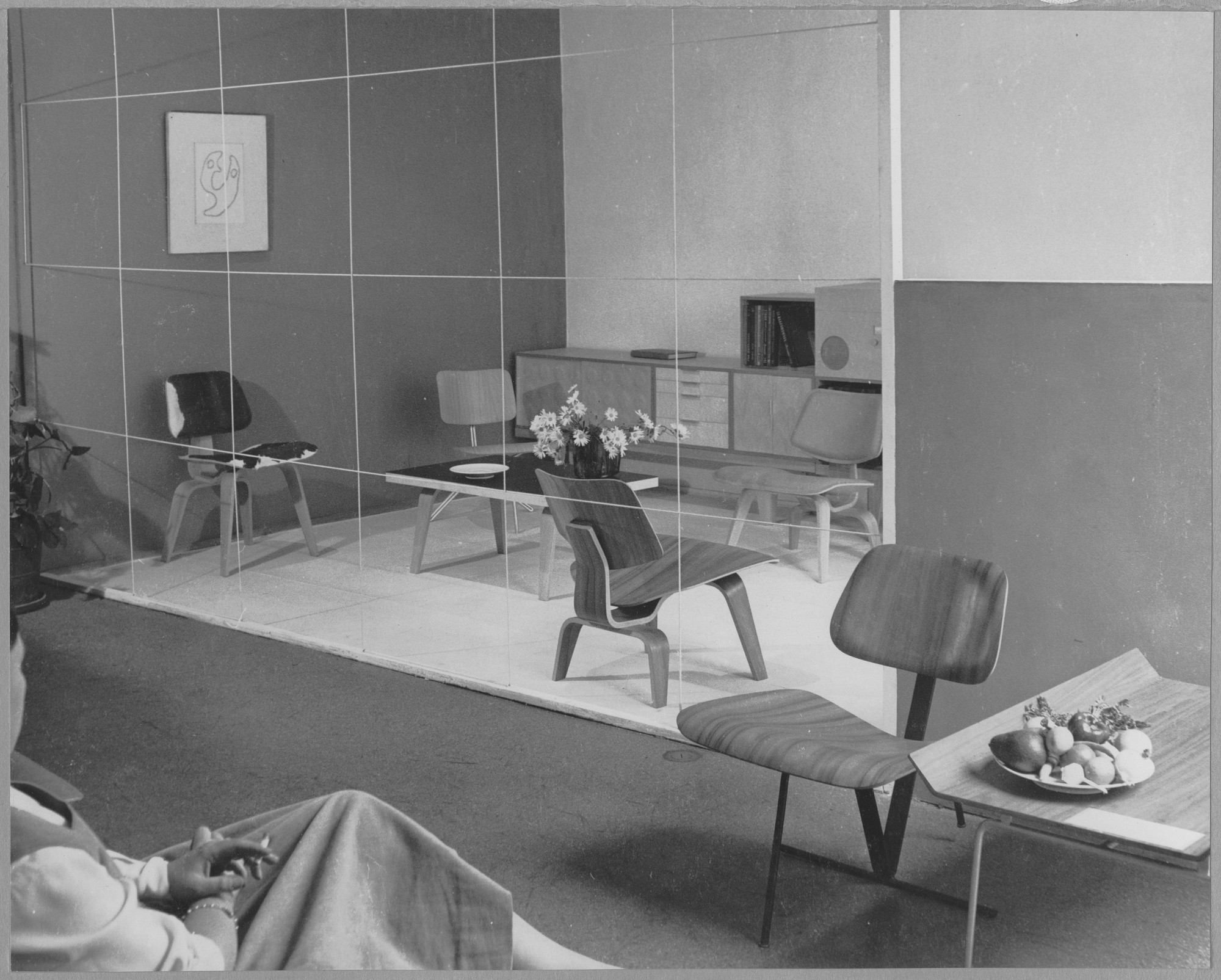 monochrome image of a room with a man seated in a chair peering through a set of window panes to a room full of chairs and a table.